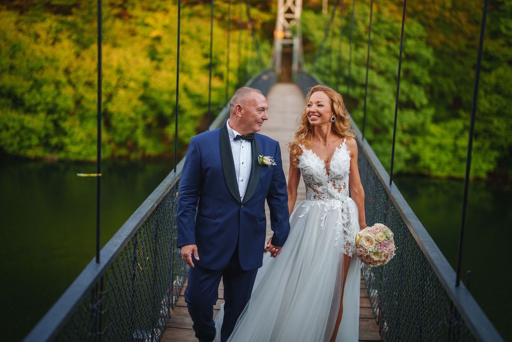 A couple in wedding attire share joyful moments while walking across a wooden bridge over a scenic lake.