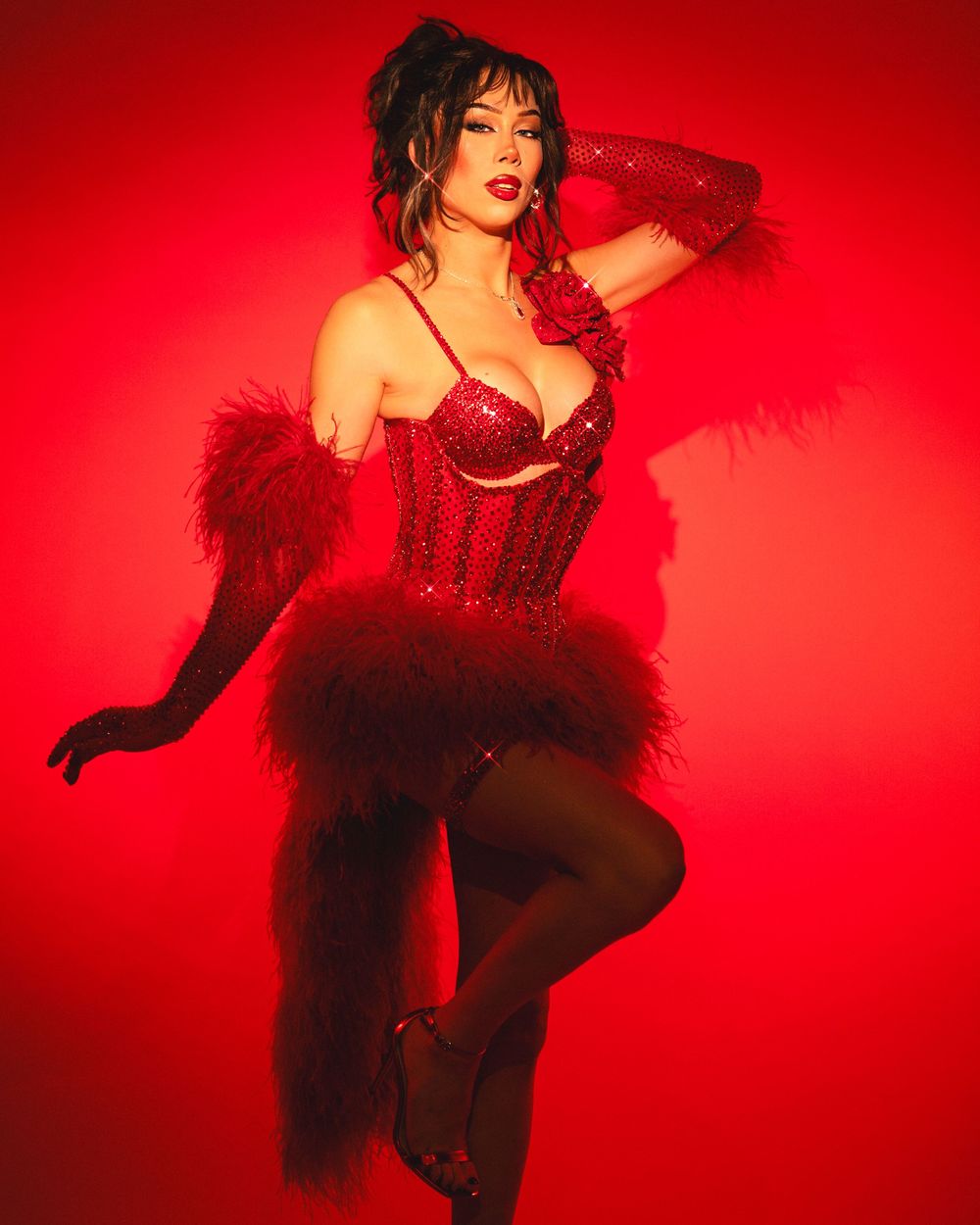 Burlesque performer in a red sequined costume and black feathers poses dramatically against a vibrant red backdrop.