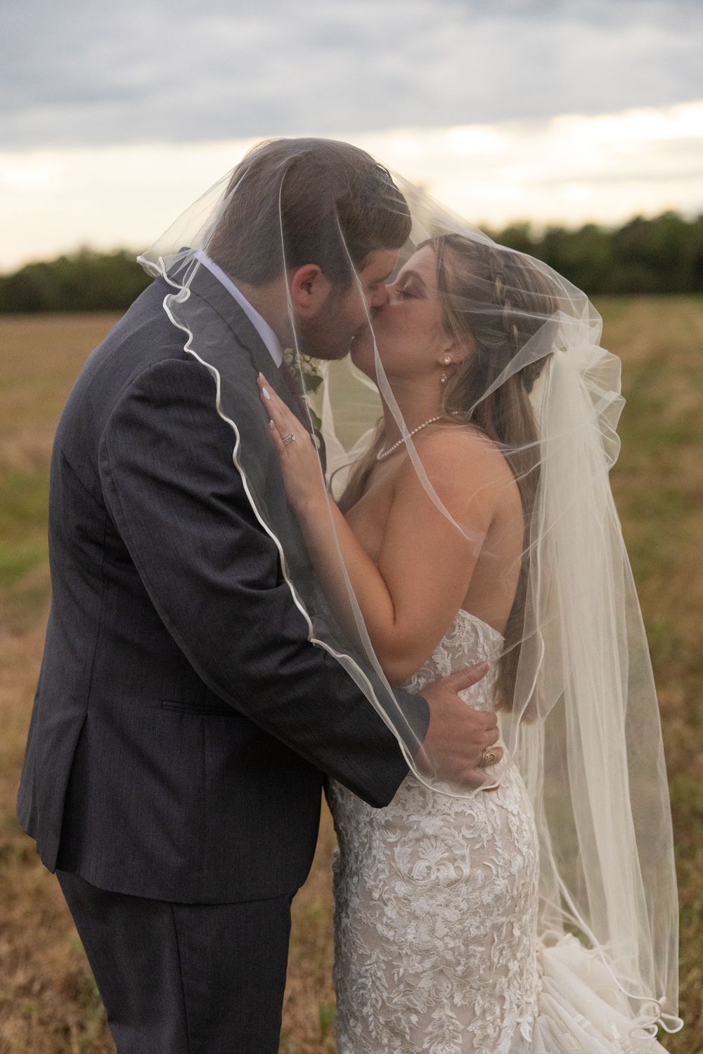 A romantic wedding couple shares intimate kisses in a golden wheat field at sunset wearing formal attire and bridal veil.
