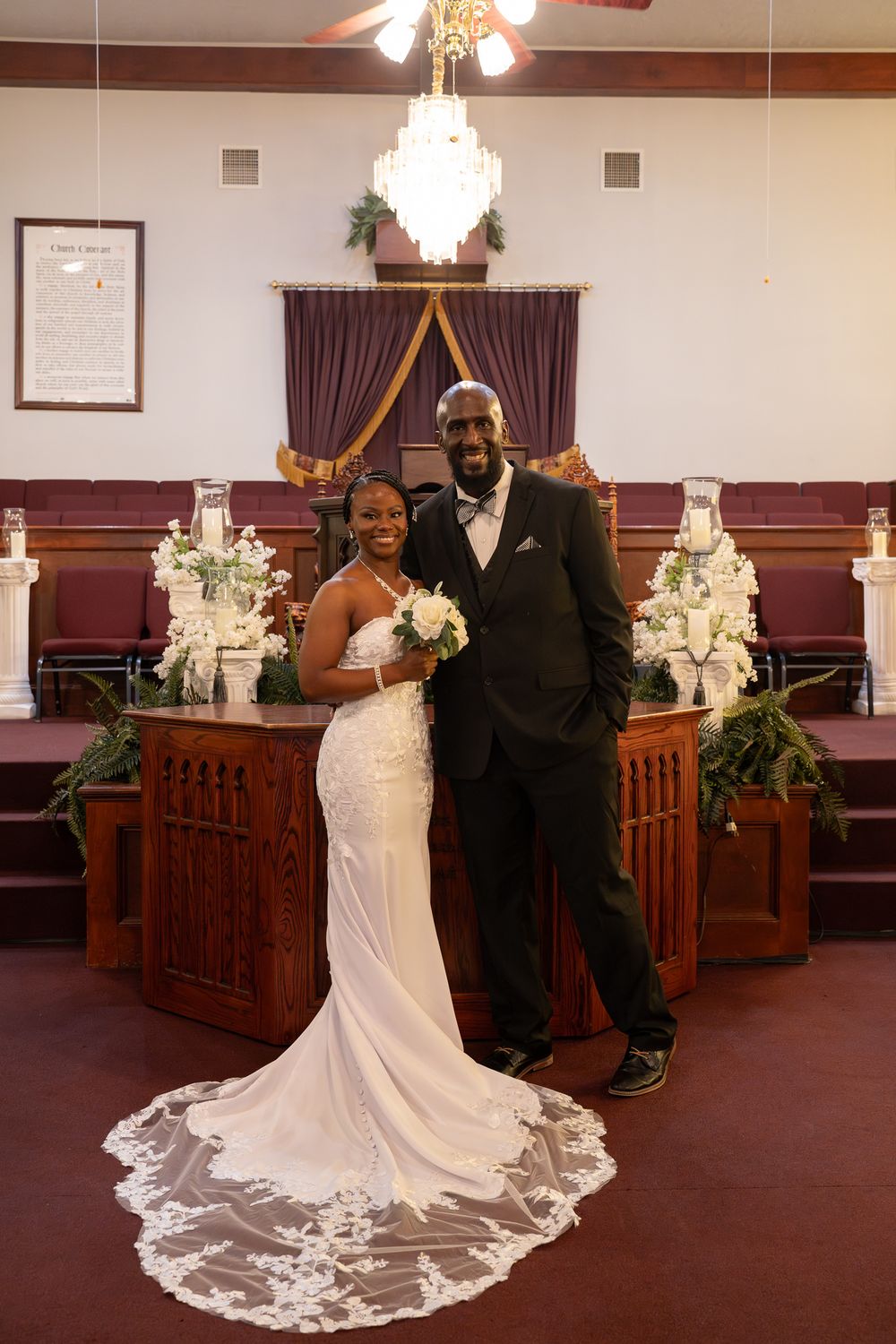 A couple poses together in a church sanctuary with elegant decorations, crystal chandeliers and wooden pews.