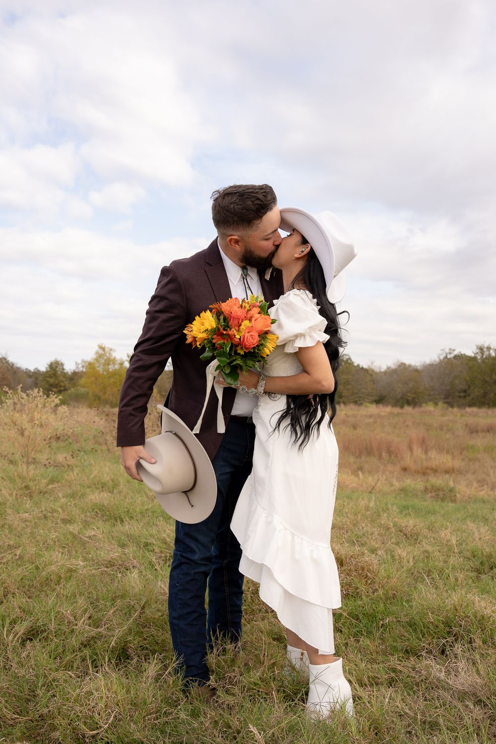 A couple shares a romantic kiss in a field while holding a vibrant autumn bouquet and cowboy hat.