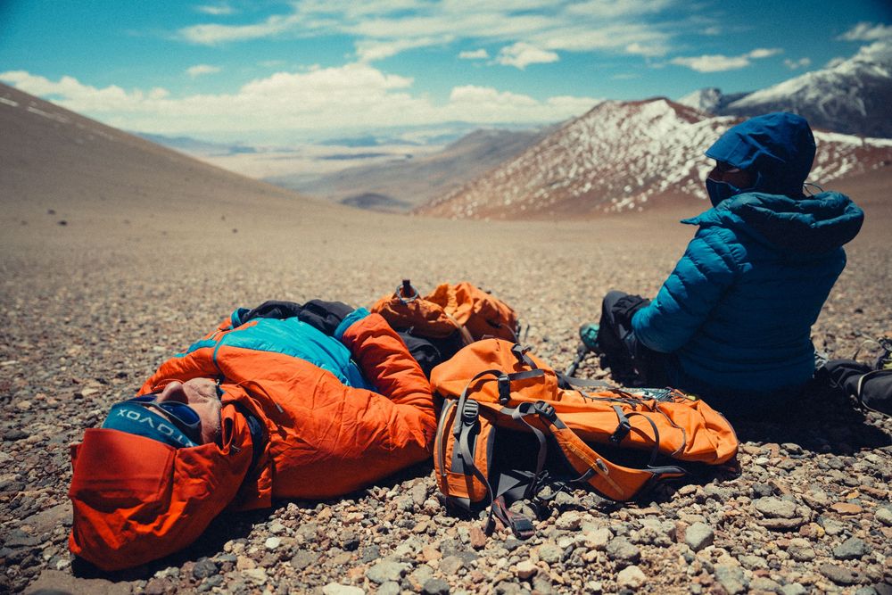 A sleeping bag in bright orange and navy blue lies on rocky desert terrain with mountains in the background.