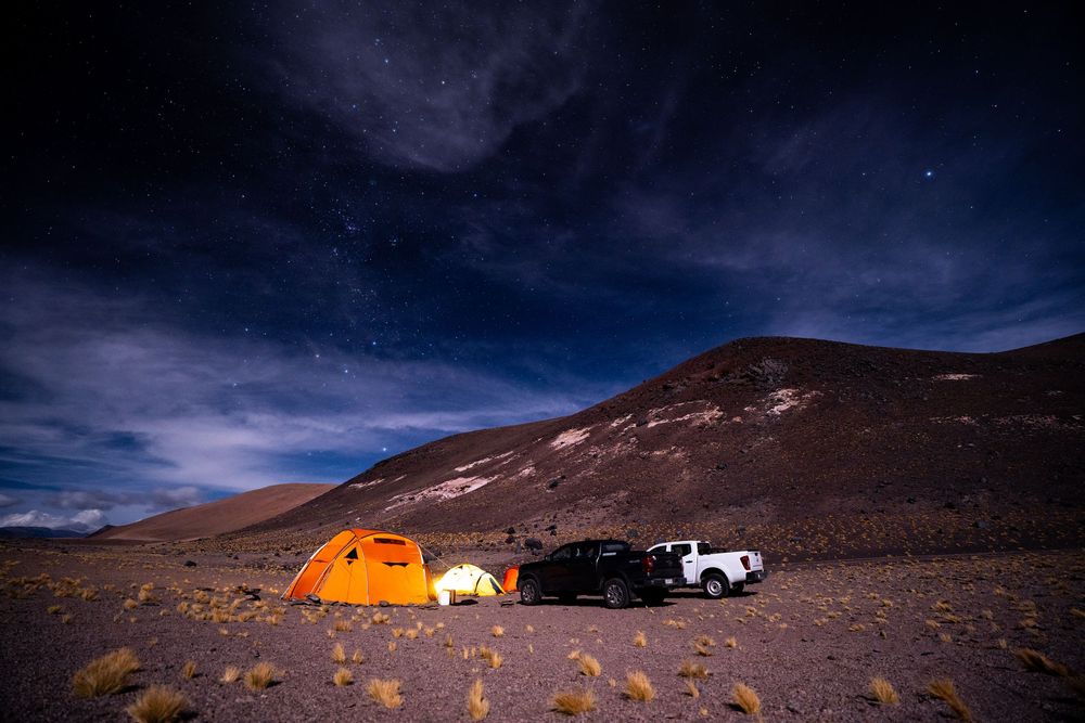 Orange tent glows under starry night sky next to parked vehicle in remote mountain desert landscape.