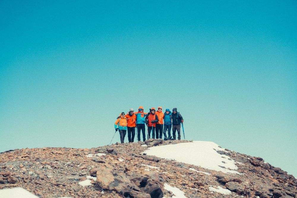 A group of hikers stands on a rocky mountain summit against a bright turquoise sky.