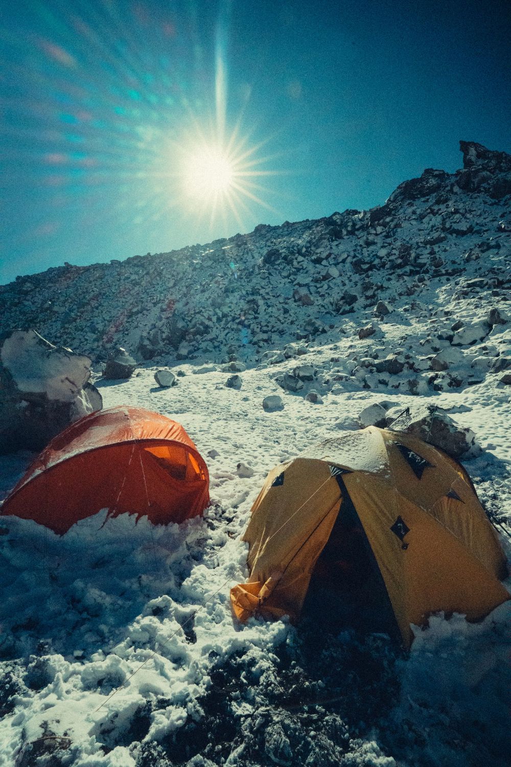 Colorful camping tents set up in snowy mountainous terrain under bright sunlight and clear blue skies.