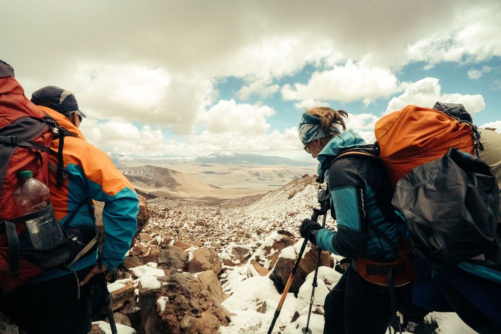 A group of hikers with backpacks traverse a rocky mountain ridge against a cloudy sky.