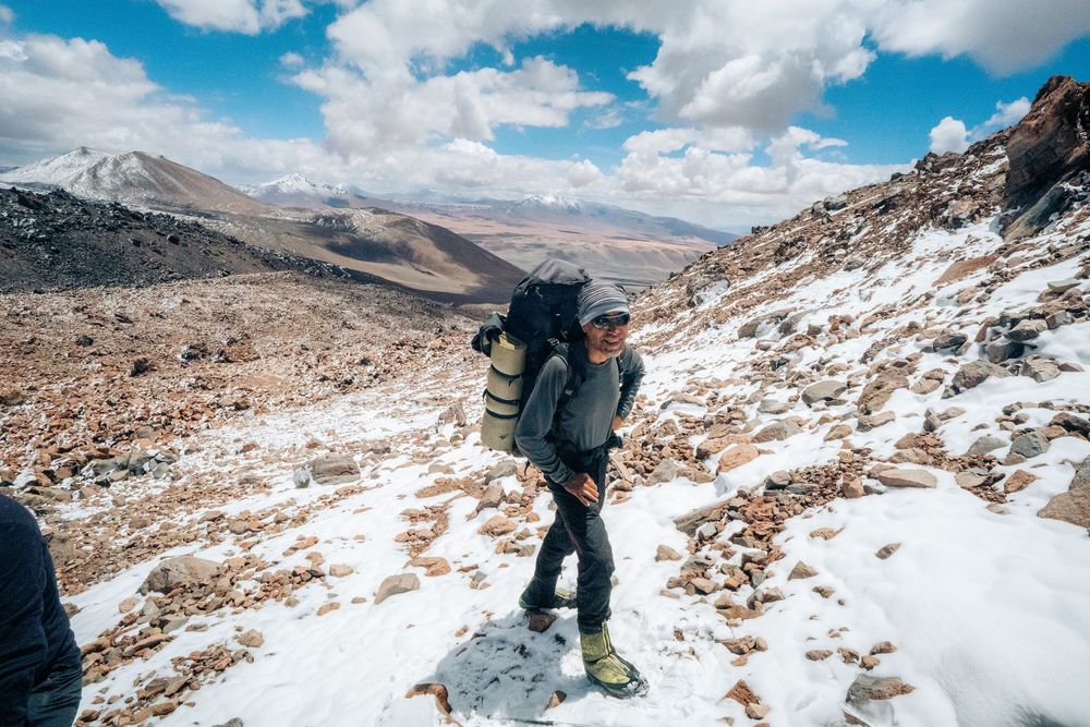 Hiker traversing snowy mountain terrain with dramatic cloudy skies and rocky peaks in the background.
