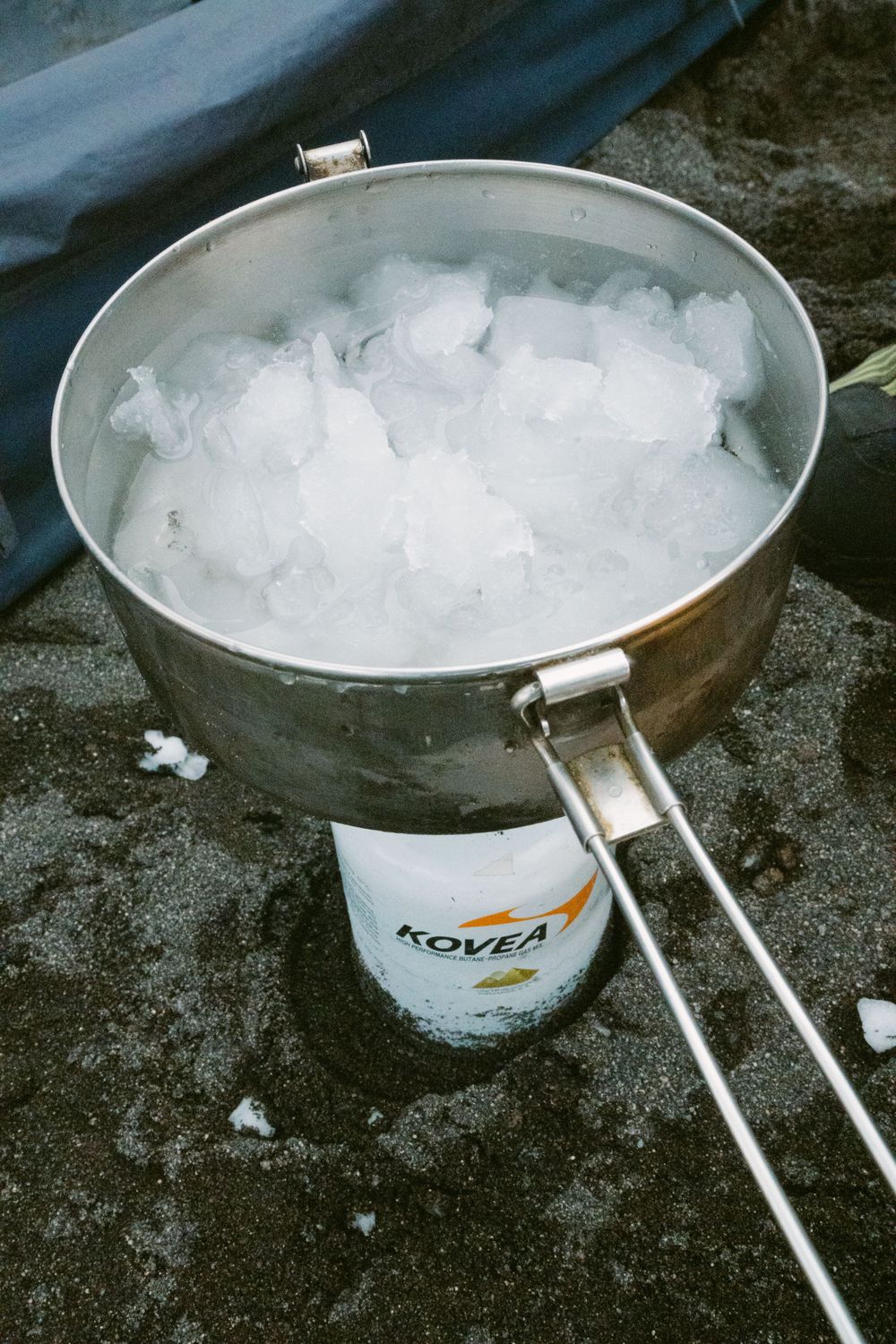 A metal camping pot filled with ice sits atop a Kovea camping stove outdoors.