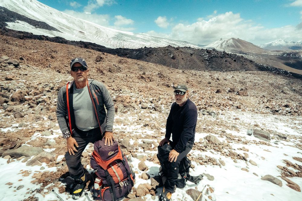 Hikers rest on a rocky mountain trail with snow-capped peaks and blue skies in the background.