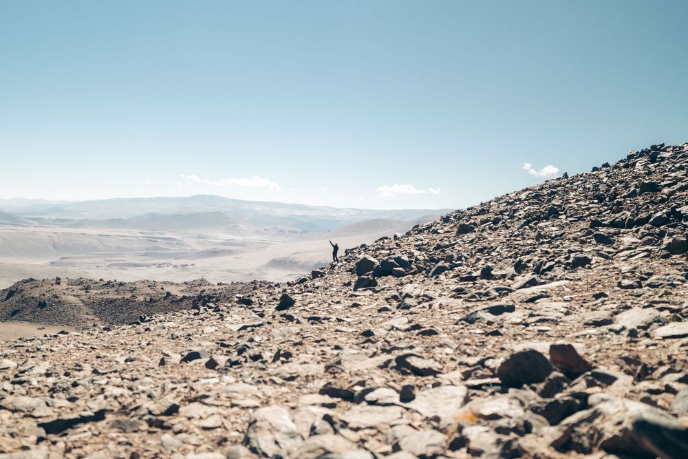 A lone hiker stands on a rocky mountainside overlooking a vast desert valley with mountains in the distance.
