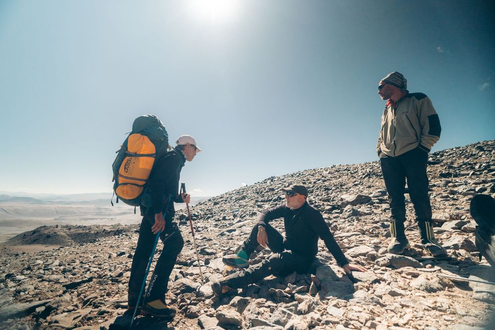 Three hikers take a break on a rocky mountain summit against a bright blue sky with backpacks and hiking gear.