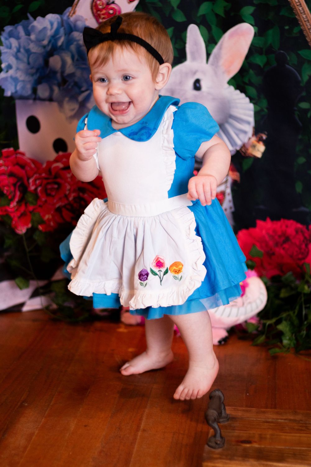 A baby in a white dress and blue wings smiles while crawling on a hardwood floor with red poinsettias in the background.