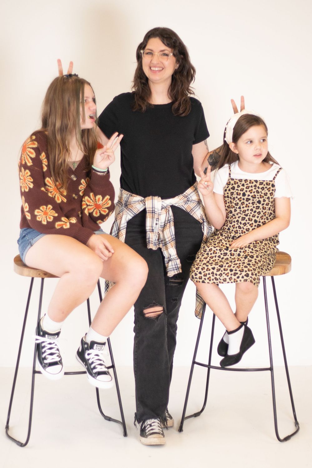 Three people pose playfully on bar stools wearing casual outfits including floral print, black, and leopard print against white backdrop.