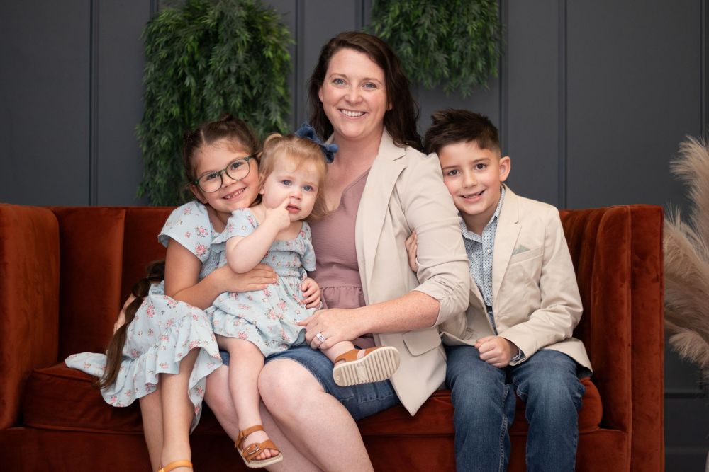 A family group sits together on a rust-colored velvet couch against a dark wall with green plants in the background.