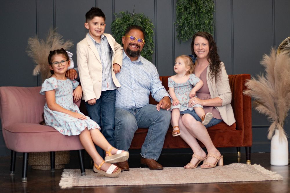 A family of five sits together on a pink couch with decorative pampas grass in the background.