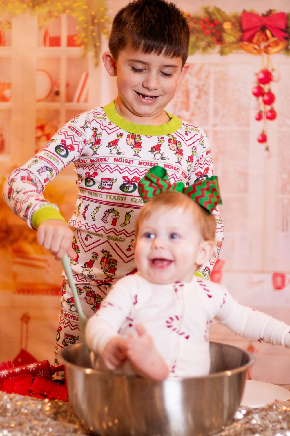 Two children in matching Christmas pajamas play together with a large metal mixing bowl during holiday baking.