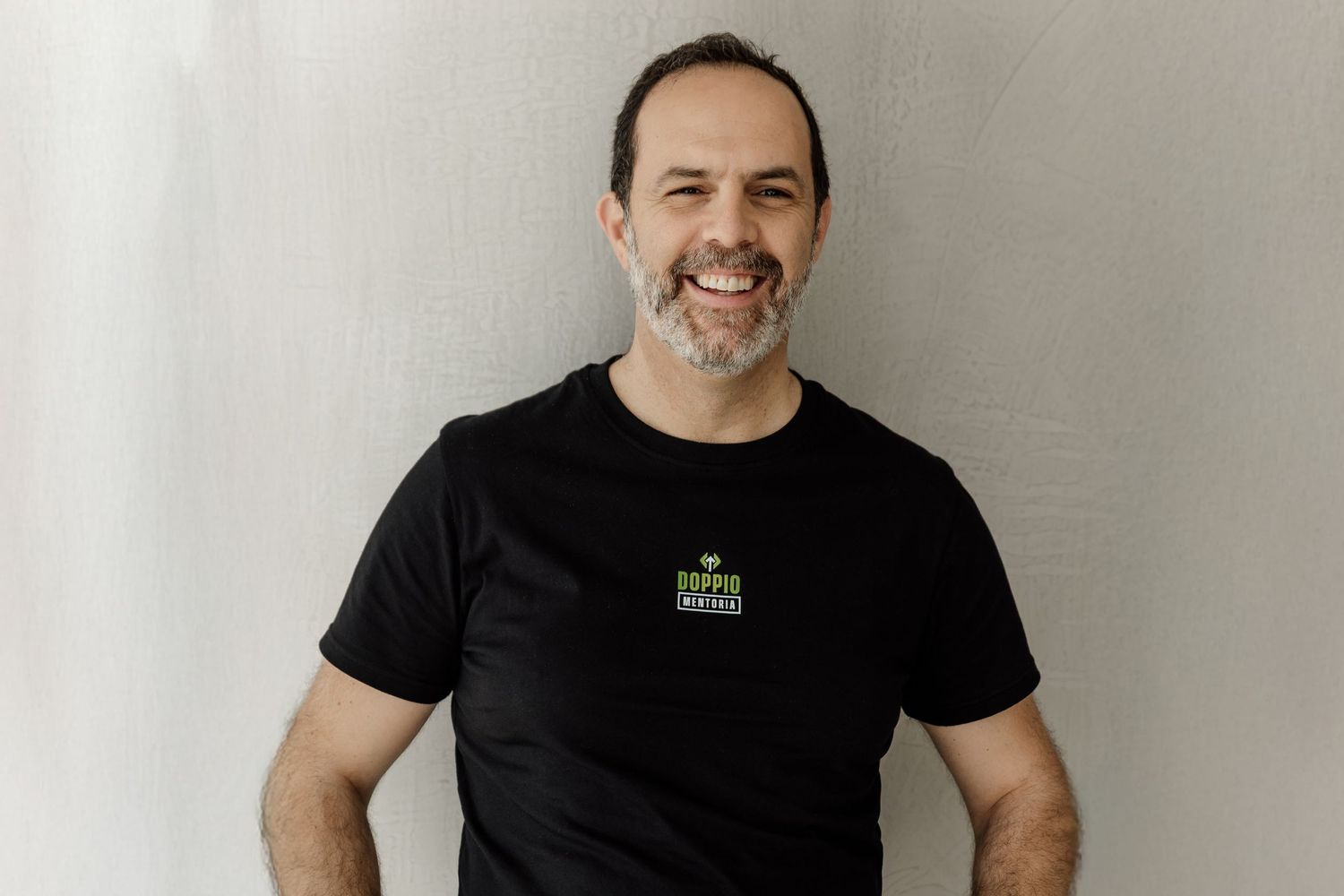 Someone wearing a black t-shirt with a small logo stands in front of a light colored wall.