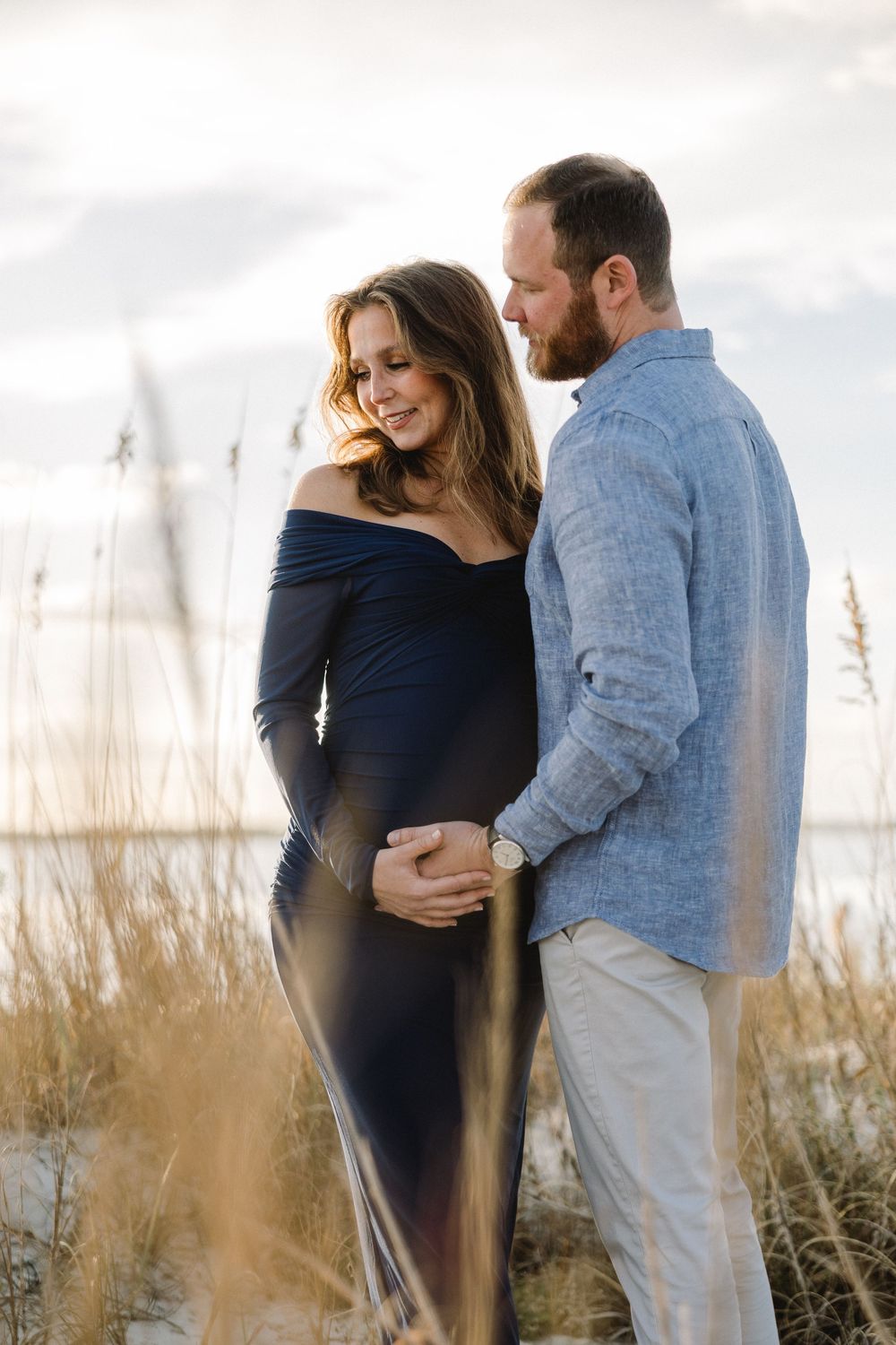A romantic couple embraces in tall beach grass during a sunset photoshoot at the shore.