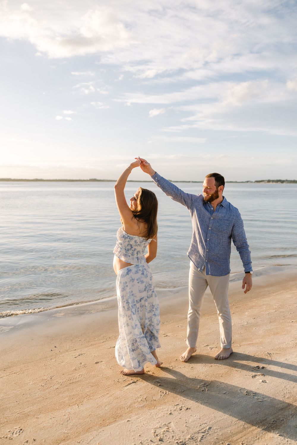 A couple dances playfully together on a sunny beach with waves and cloudy sky in the background.