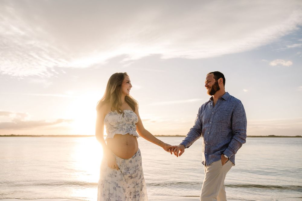 A couple holds hands on a beach at sunset with golden sunlight streaming between them over calm ocean waters.