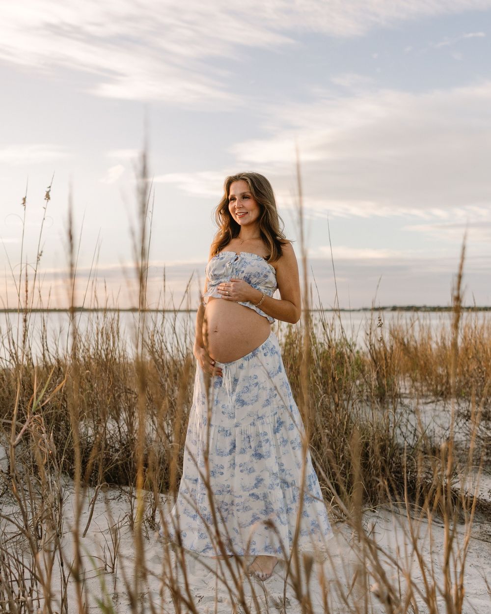Maternity photo session among tall beach grass with coastal waters and cloudy sky in the background.
