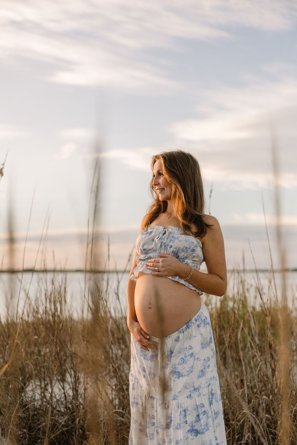 Expectant mother in a floral dress poses against tall grass and cloudy sky during sunset maternity photoshoot.
