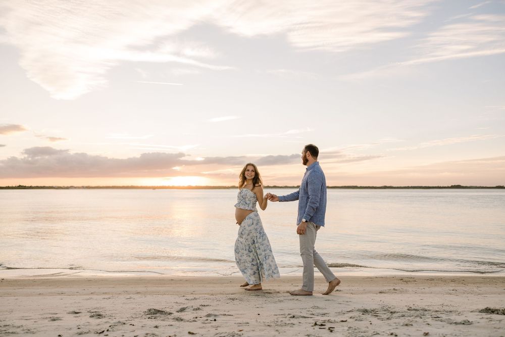 A couple walks along a serene beach at sunset with warm golden light reflecting on the sand and water.