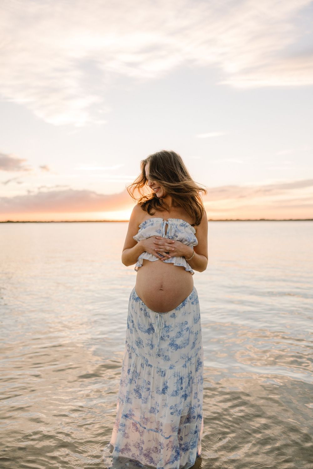 Pregnant woman in floral blue dress stands by calm waterfront during golden hour sunset.