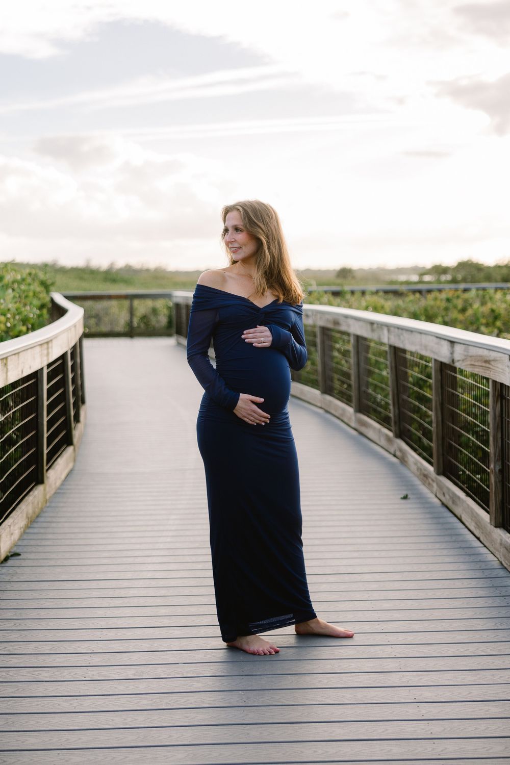 Pregnant woman in navy dress sits thoughtfully on wooden boardwalk overlooking coastal wetlands.