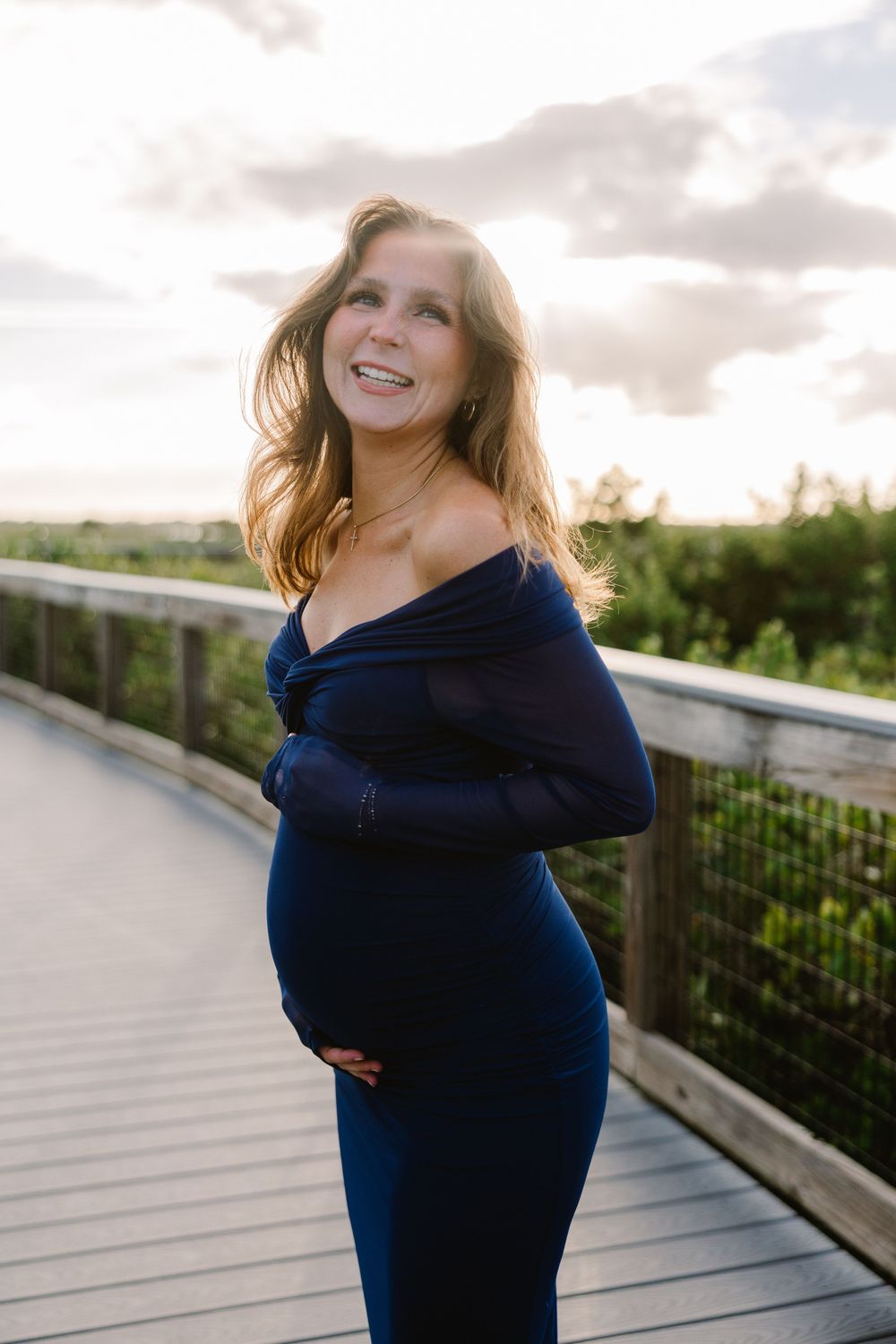 Maternity photo of woman in navy dress twirling on wooden boardwalk at sunset.