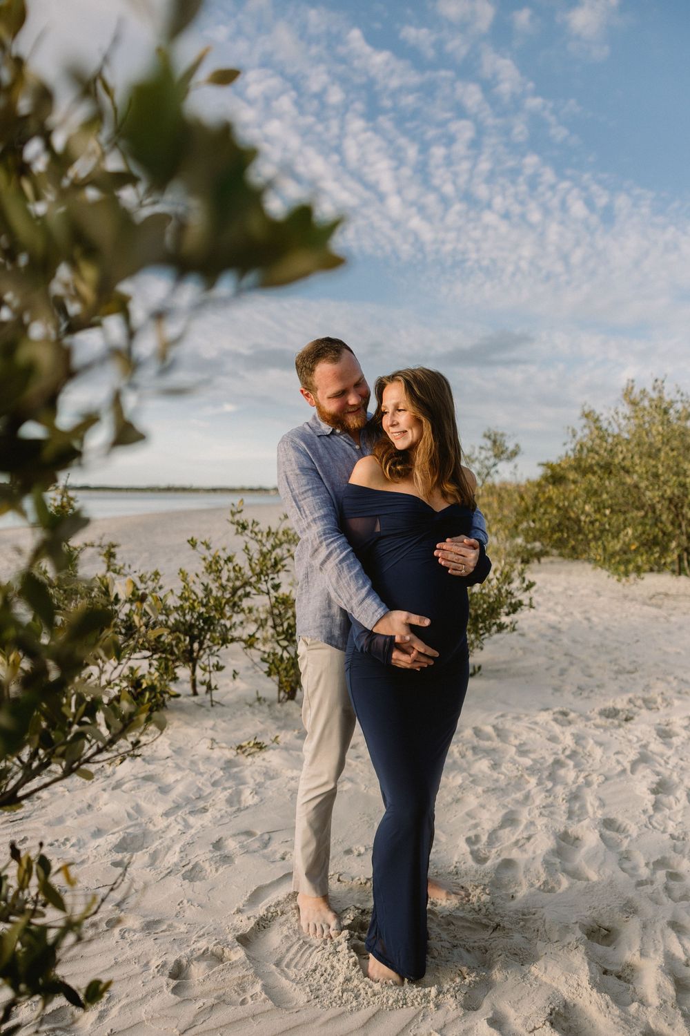 Romantic couple embraces on sandy beach with scattered clouds and coastal vegetation.