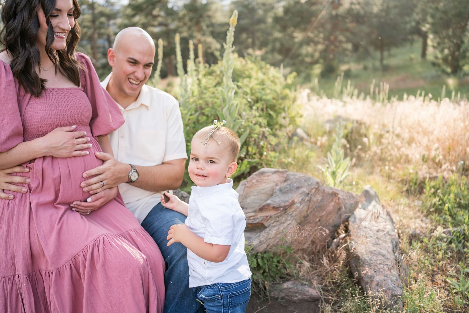 Family maternity photo outdoors with toddler in white shirt and mother in pink dress near garden rocks.
