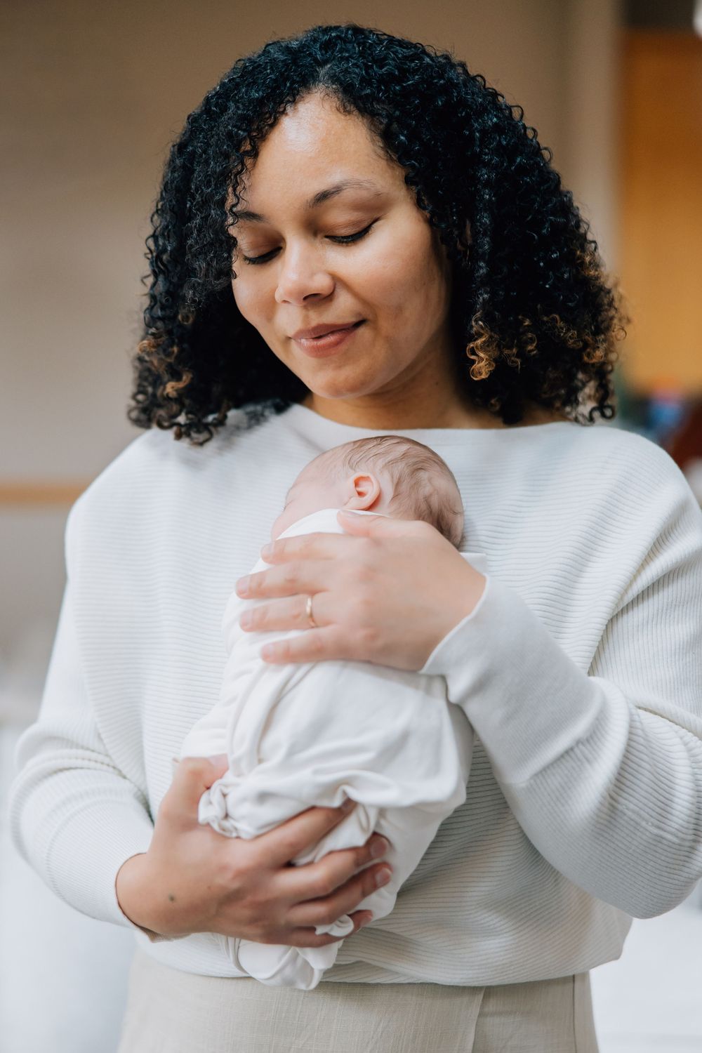 Smiling curly-haired woman tenderly holding swaddled newborn baby.