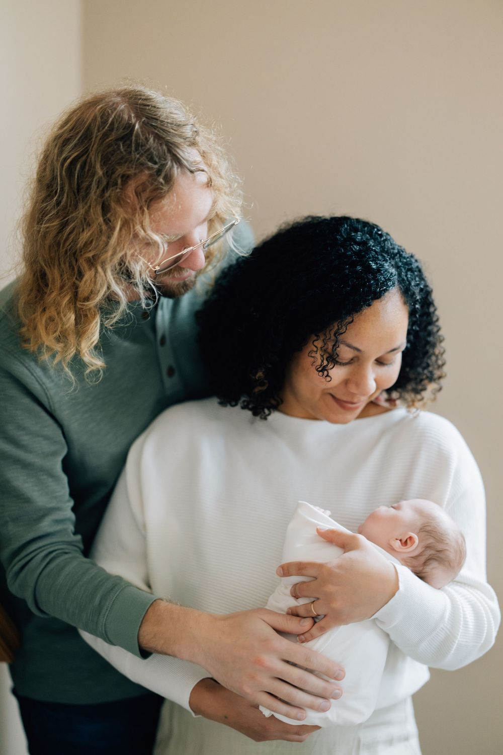 Interracial couple lovingly gazing at their swaddled newborn baby together.