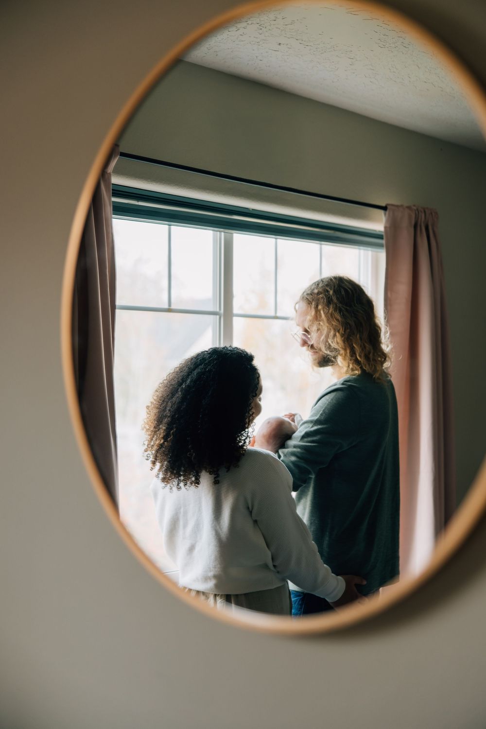 Couple with newborn baby reflected in round mirror by window.