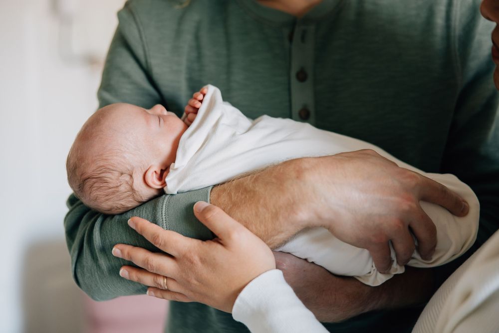 Father cradling sleeping newborn baby wrapped in white swaddle blanket.