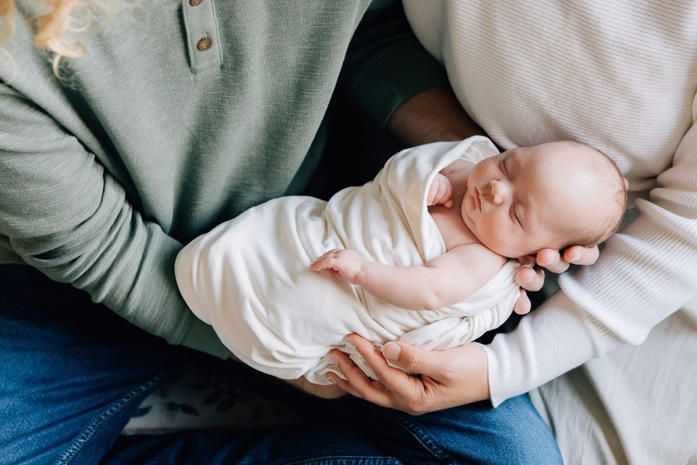 Parents holding sleeping newborn baby dressed in white outfit.
