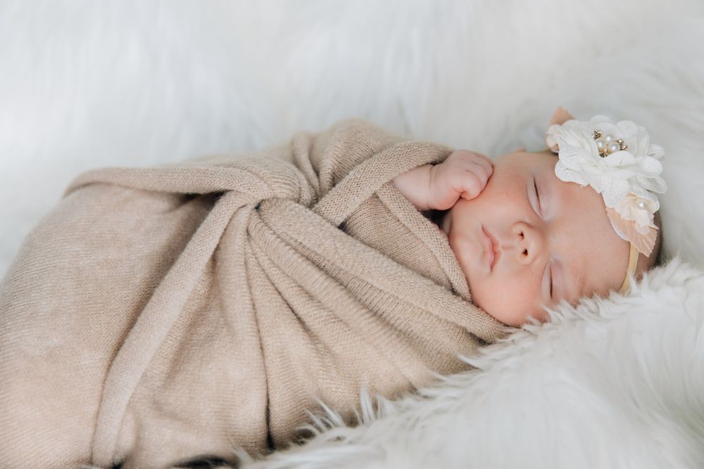 Sleeping newborn baby swaddled in beige blanket with white floral headband.
