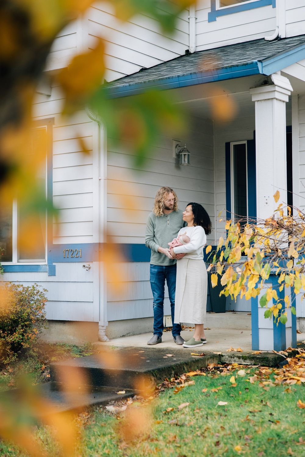 Interracial couple with newborn baby standing on front porch in autumn.