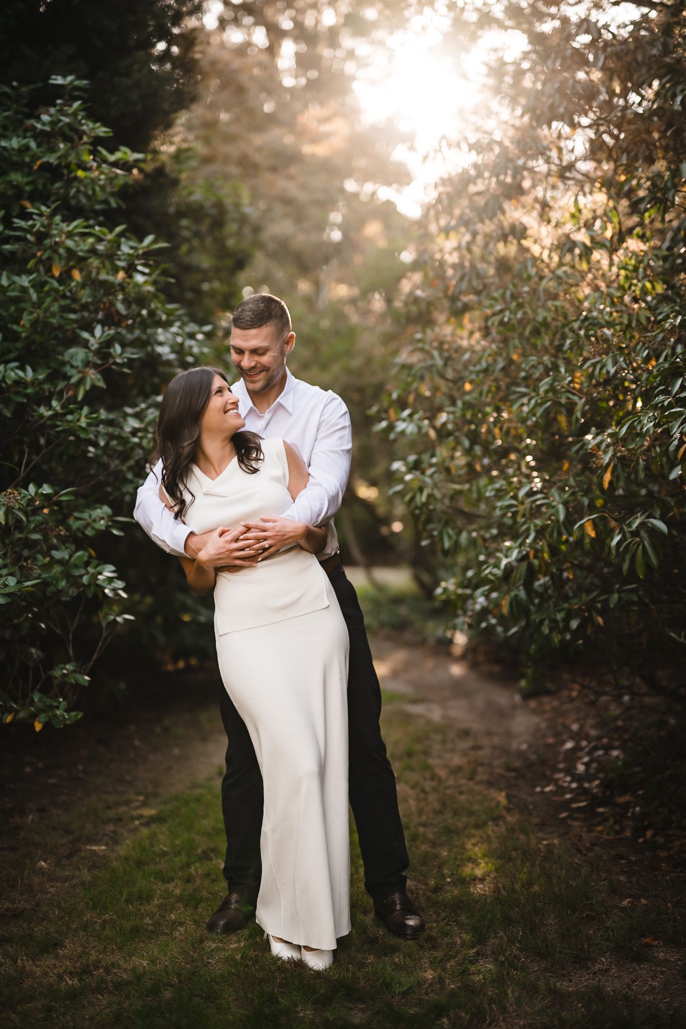 Couple in white and black formal wear embracing in a sunlit garden setting.