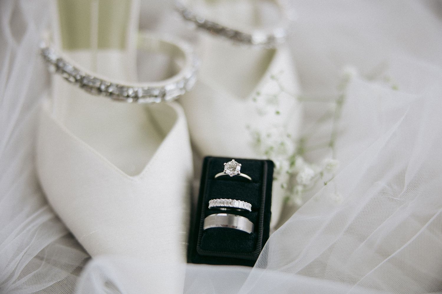 Wedding rings and white bridal shoes with crystal details displayed on tulle fabric.