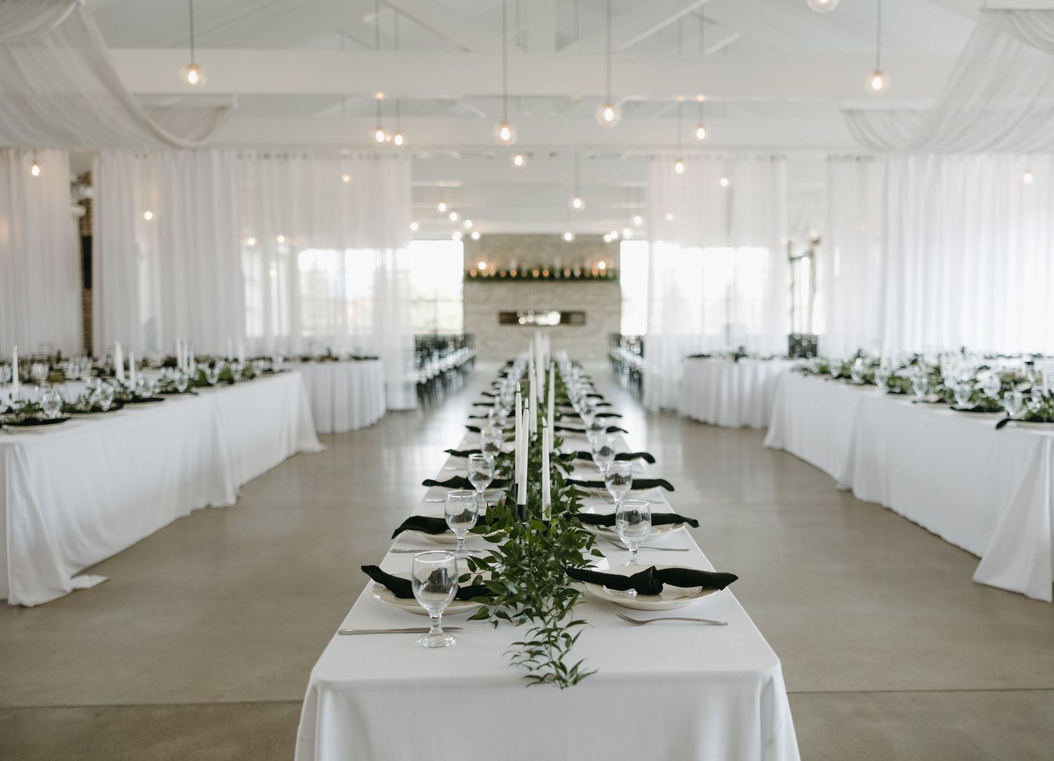 Long wedding reception table with white linens and minimal greenery in bright modern venue.