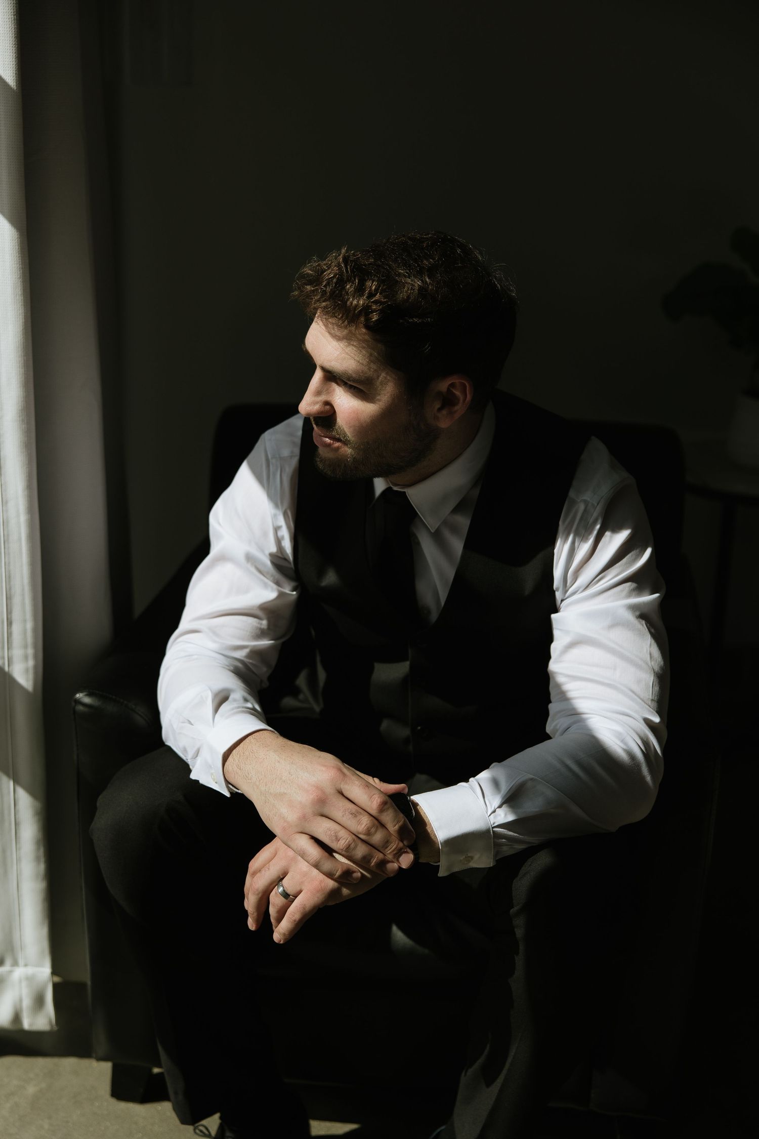 Groom in formal vest gazes thoughtfully through window in moody lighting.
