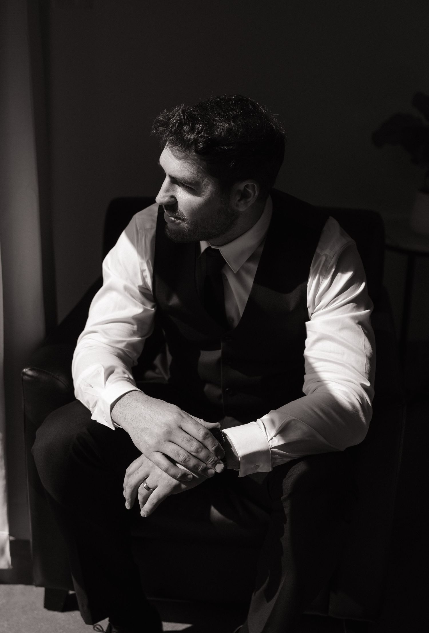 A moody black and white portrait of a well-dressed man in a vest sitting pensively in low light.