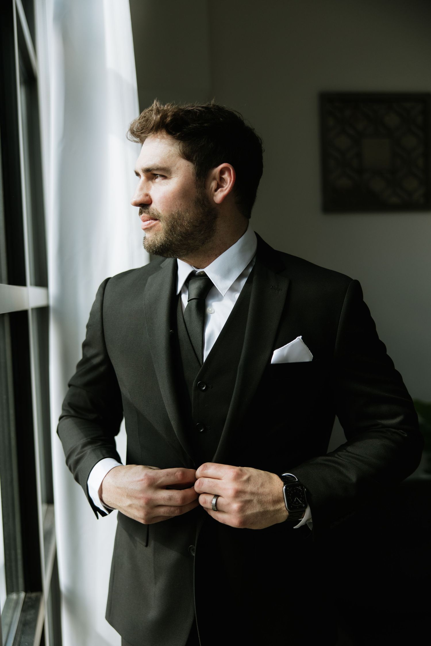 A groom in a dark three-piece suit and tie stands by a window, adjusting his cuffs.