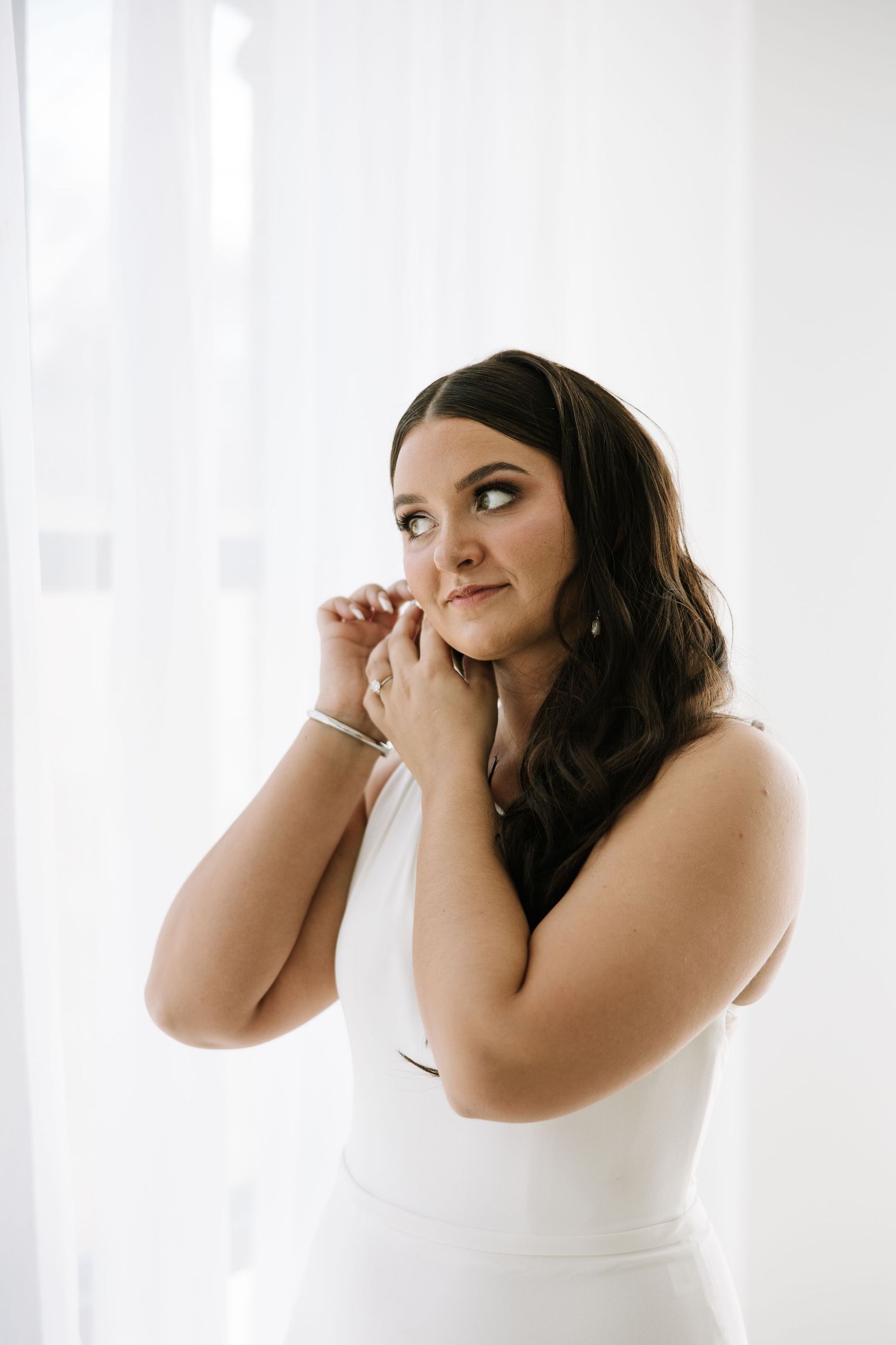 A bride wearing a white sleeveless dress puts on earrings while getting ready.