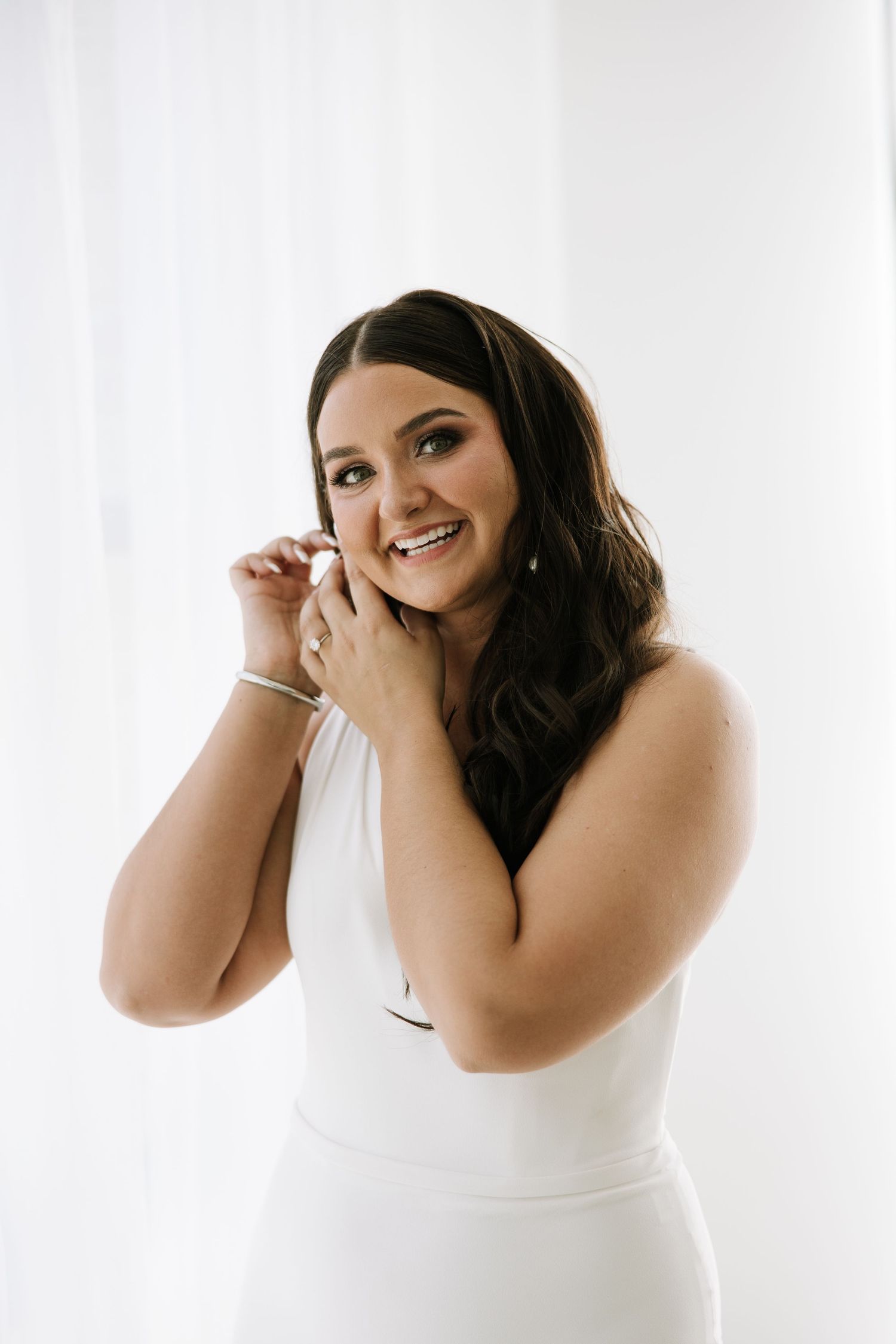 A bride in a white sleeveless dress smiles while putting on jewelry.