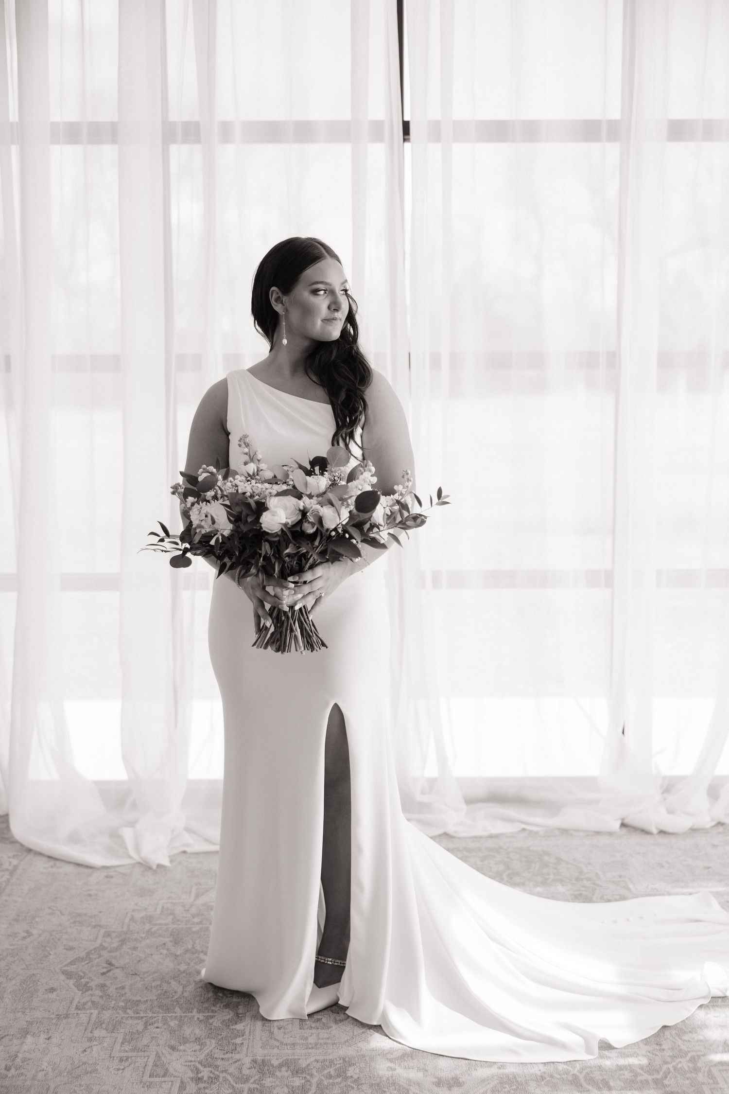 A bride in a sleek white gown holds a beautiful bouquet of flowers by a window.