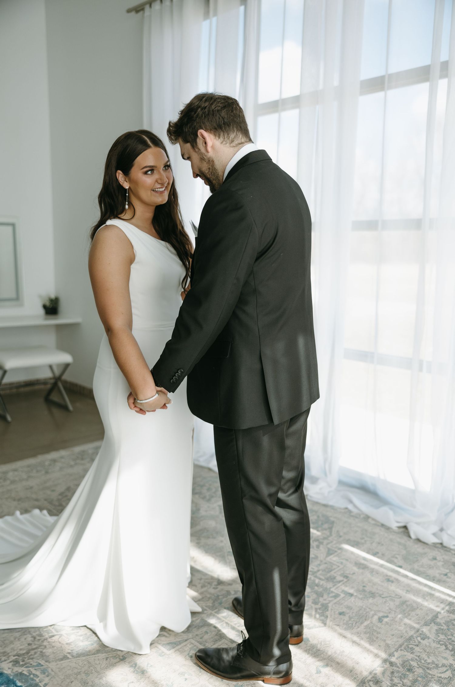 A couple in wedding attire share a joyful moment together by a bright window.