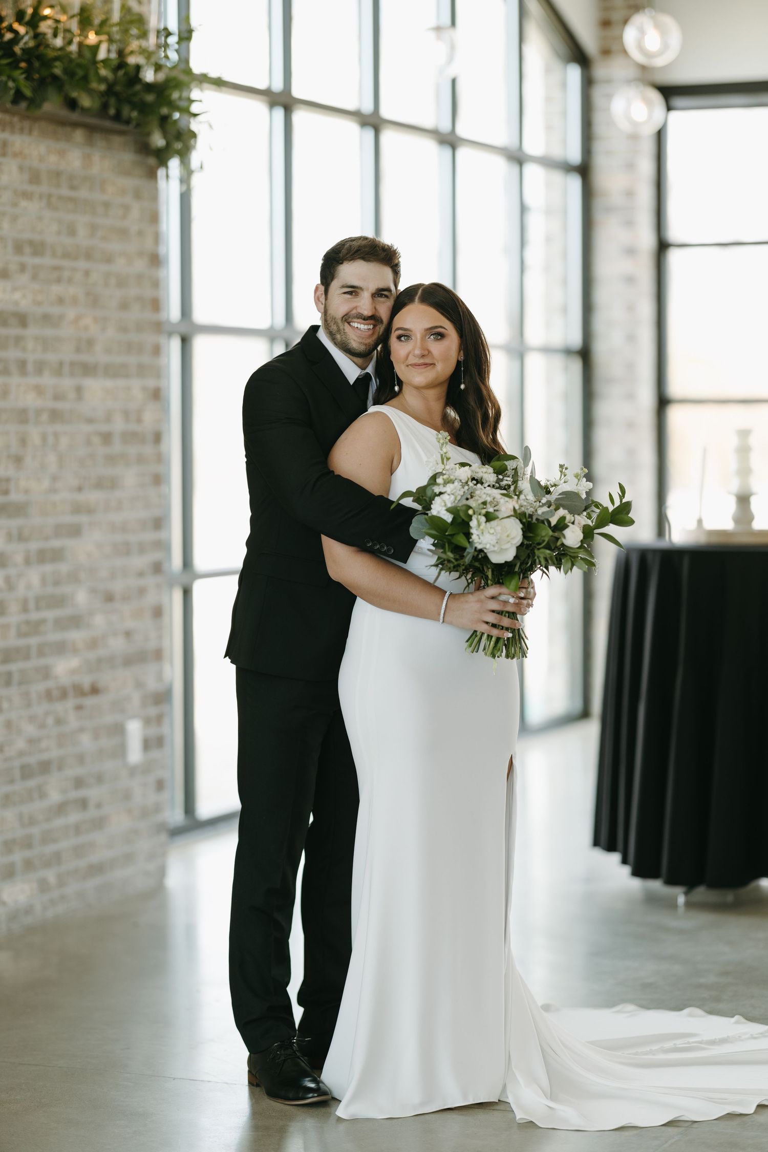 An elegant couple poses in formal wedding attire against a modern industrial brick and window backdrop.